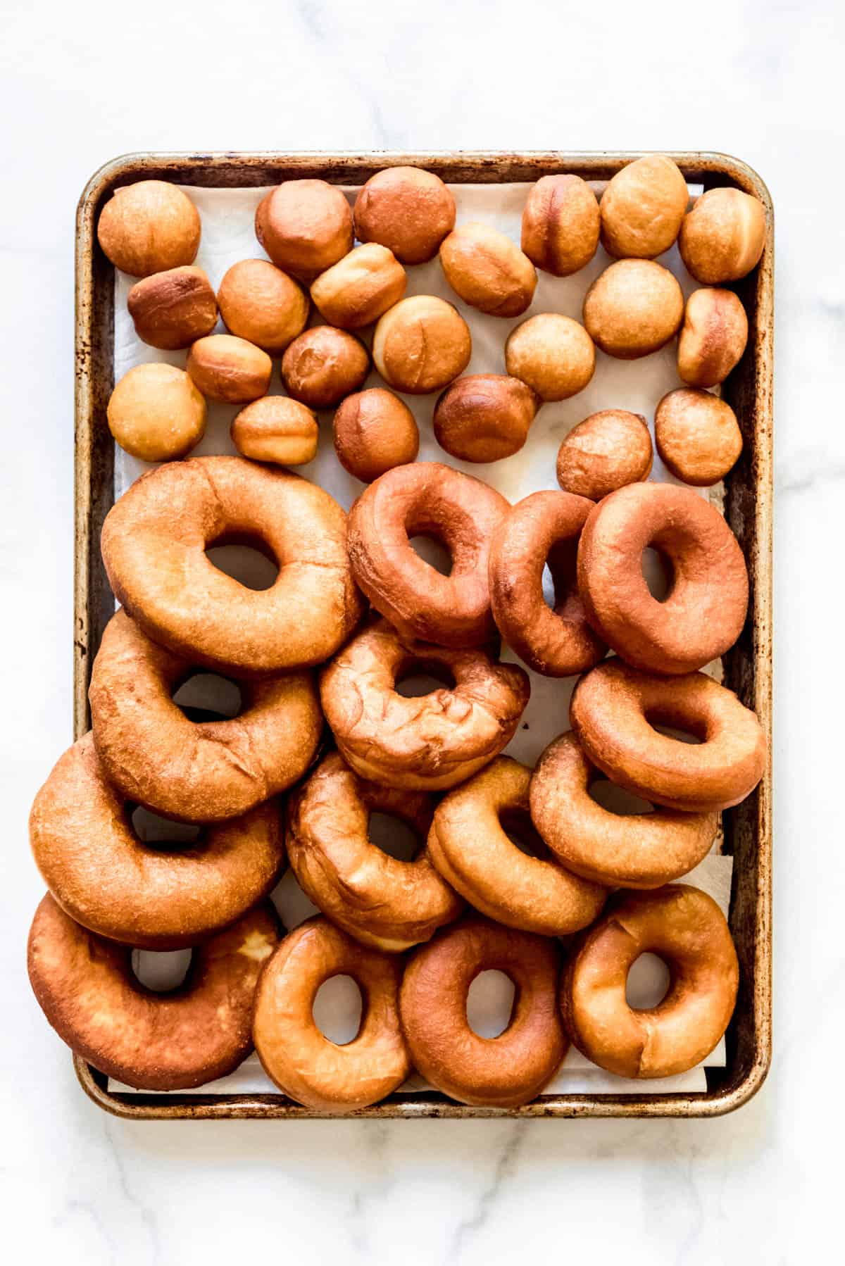 An overhead image of freshly fried hot donuts and donut holes on a baking sheet.