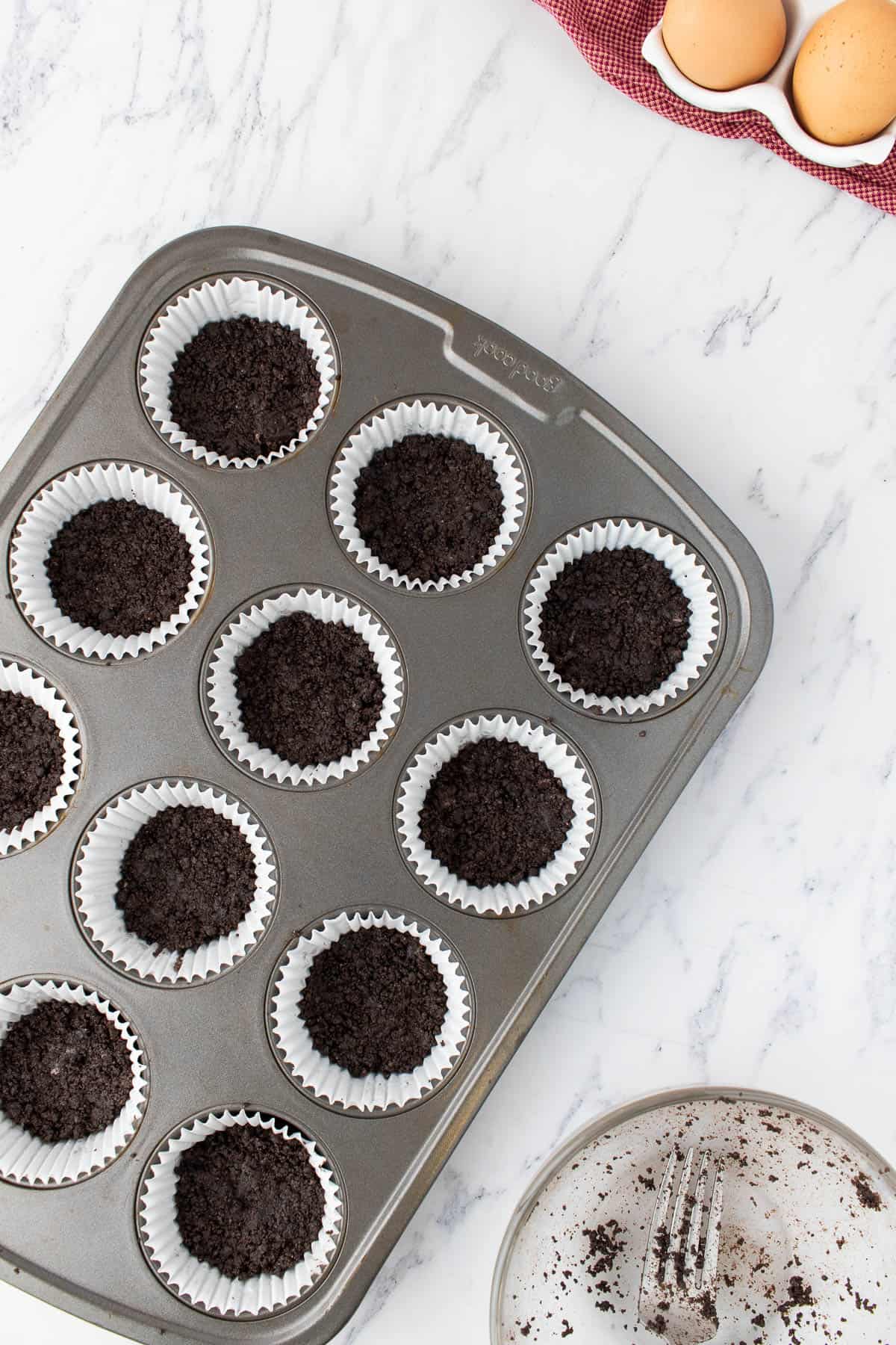 Adding crushed Oreo crust to muffin liners in a cupcake pan.