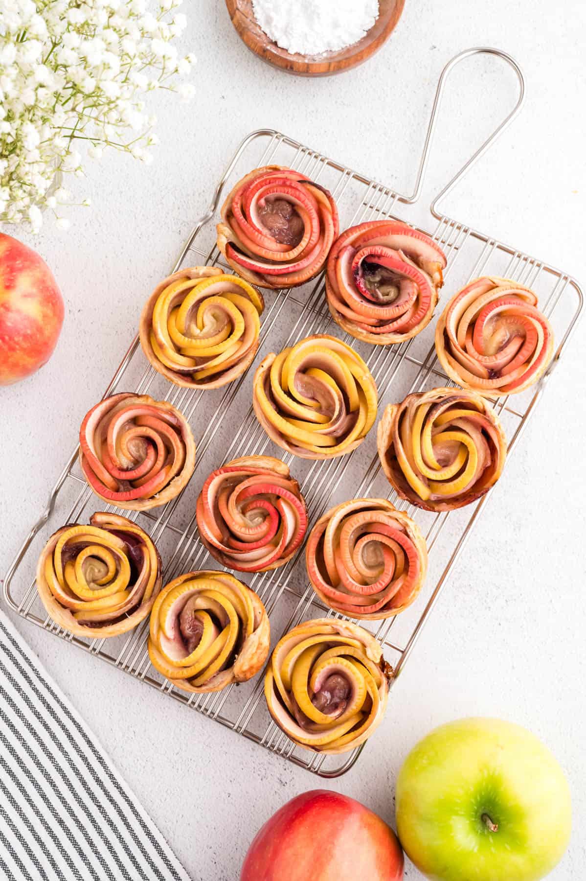 An overhead image of puff pastry apple roses on a silver cooling rack with red and green apples around it.