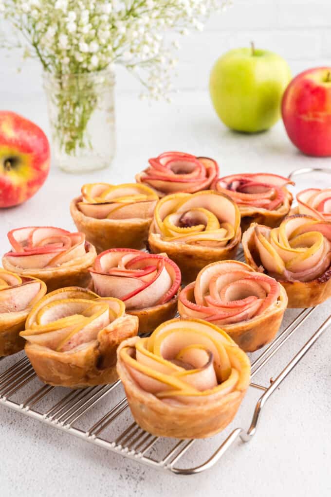 Red and yellow puff pastry apple rose desserts on a wire rack in front of red and green apples.