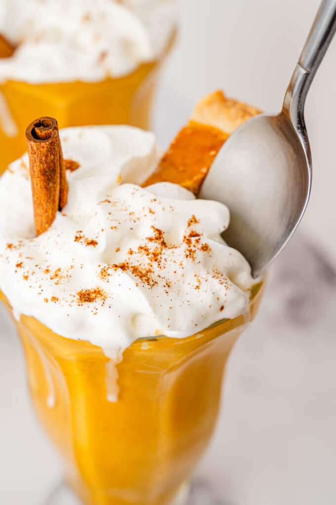 Garnishing pumpkin milkshakes with whipped cream, a cinnamon stick, and a small piece of pumpkin pie.