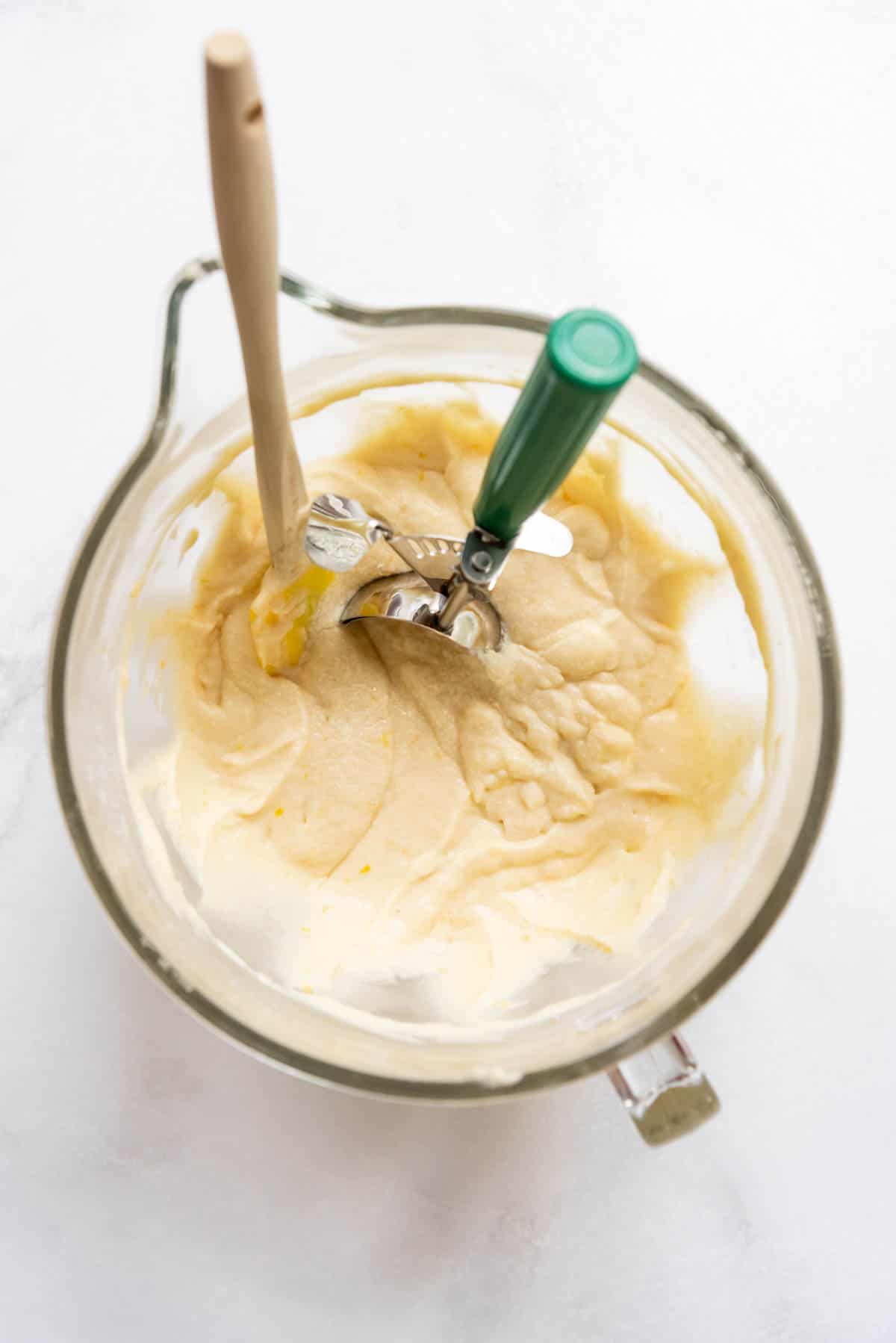 Top view of a glass mixing jug with lemon cupcake batter, a wooden spoon, and a scoop in it.
