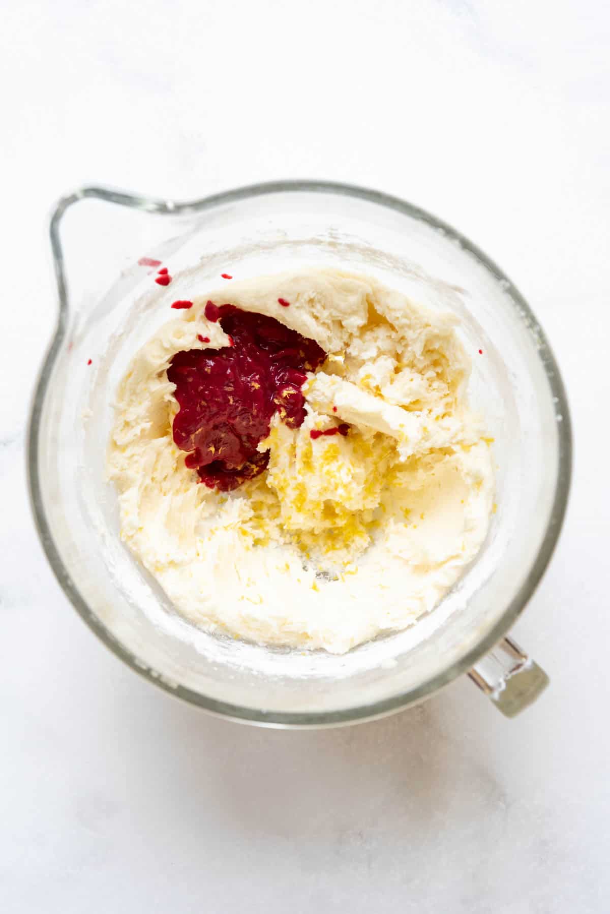 Top view of glass mixing jug with butter, vanilla, powdered sugar, lemon zest, and reduced raspberry puree in it.