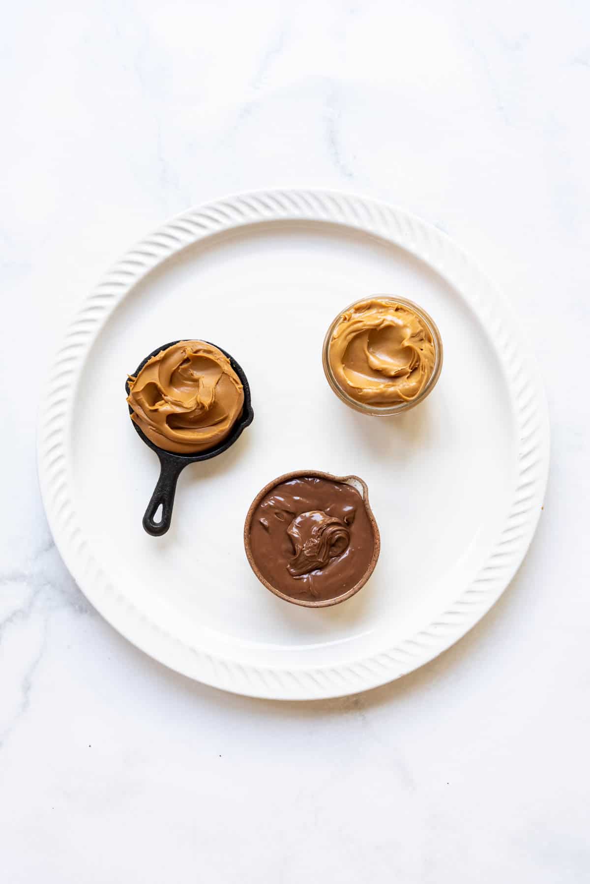 Small bowls of peanut butter, nutella, and cookie butter on a white platter.