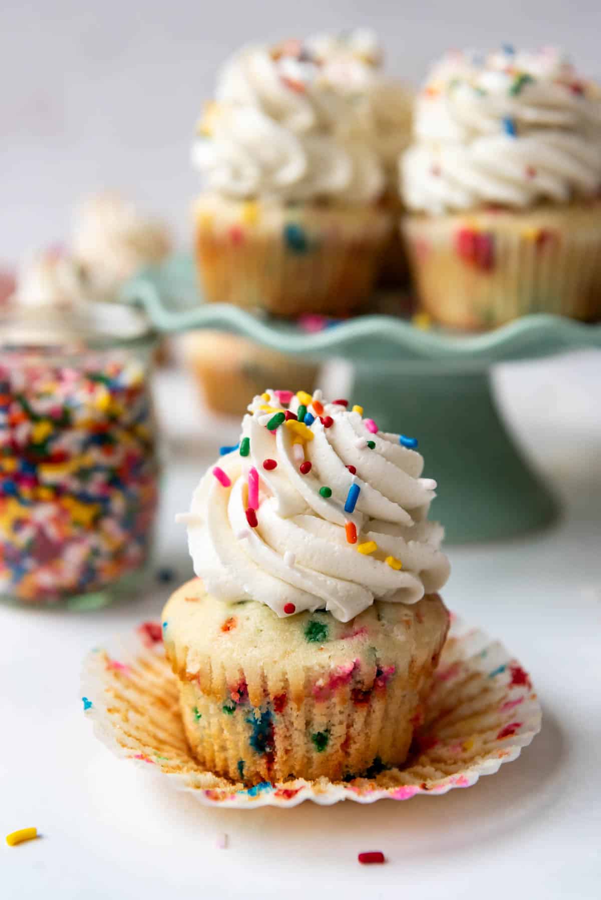 A frosted funfetti cupcake with sprinkles on top in front of more cupcakes on a pale green cake stand.