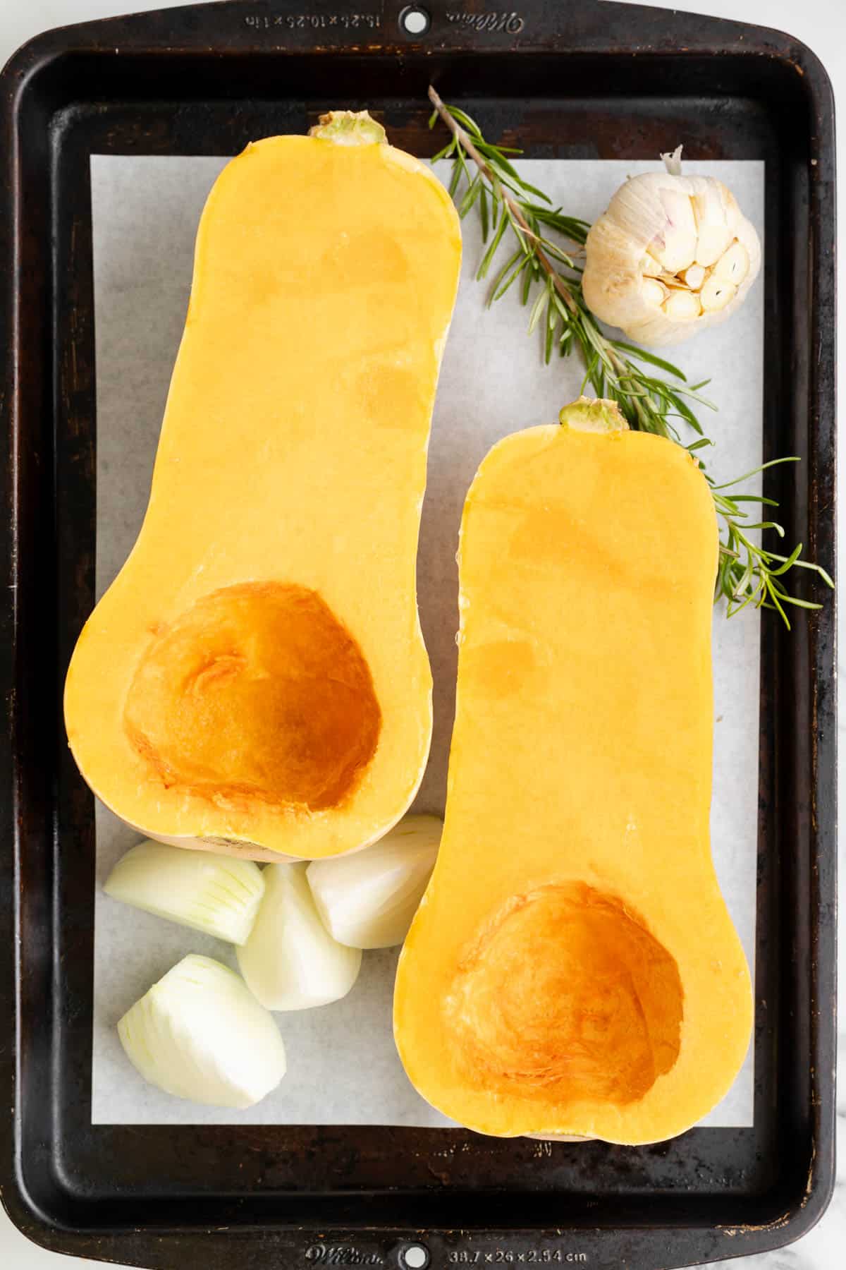 A butternut squash that has been cut in half on a pan with onion, garlic, and rosemary.