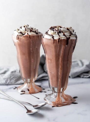 Two tall milkshake glasses filled with chocolate milkshakes and topped with whipped cream and chocolate sauce.