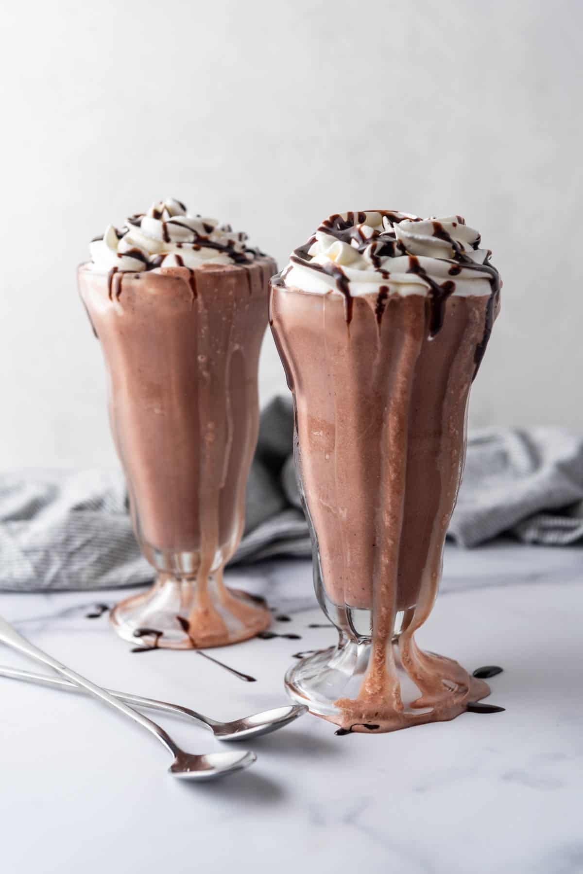Two tall milkshake glasses filled with chocolate milkshakes and topped with whipped cream and chocolate sauce.