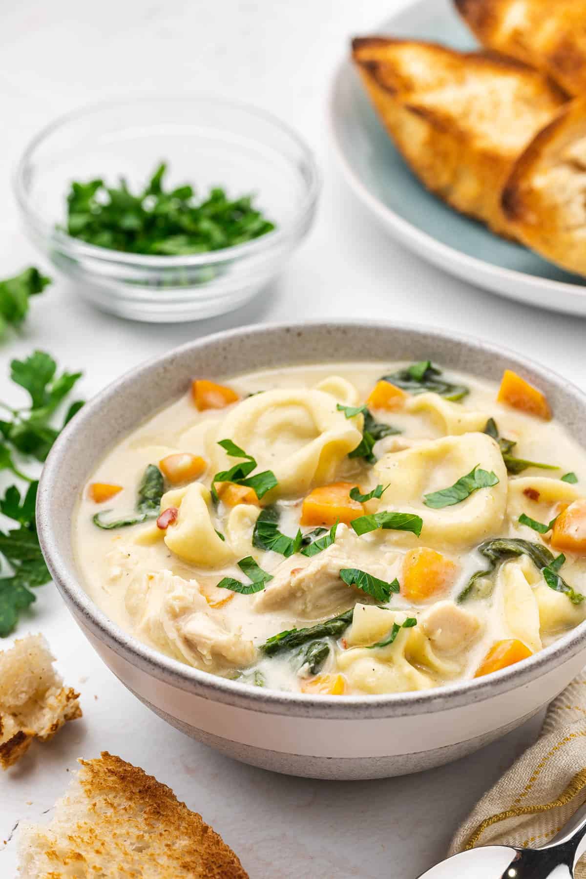A bowl of creamy chicken tortellini soup next to crusty bread and chopped fresh parsley.