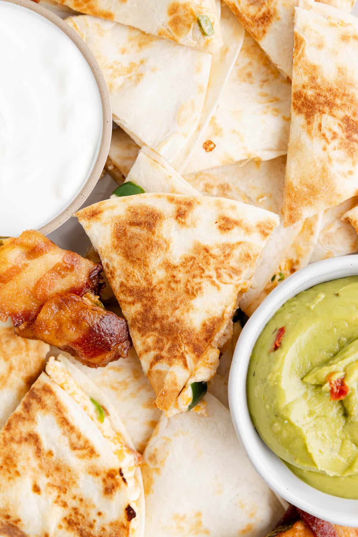 A close image of a triangle of jalapeno popper quesadilla on top of more quesadillas next to bowls of sour cream and guacamole.