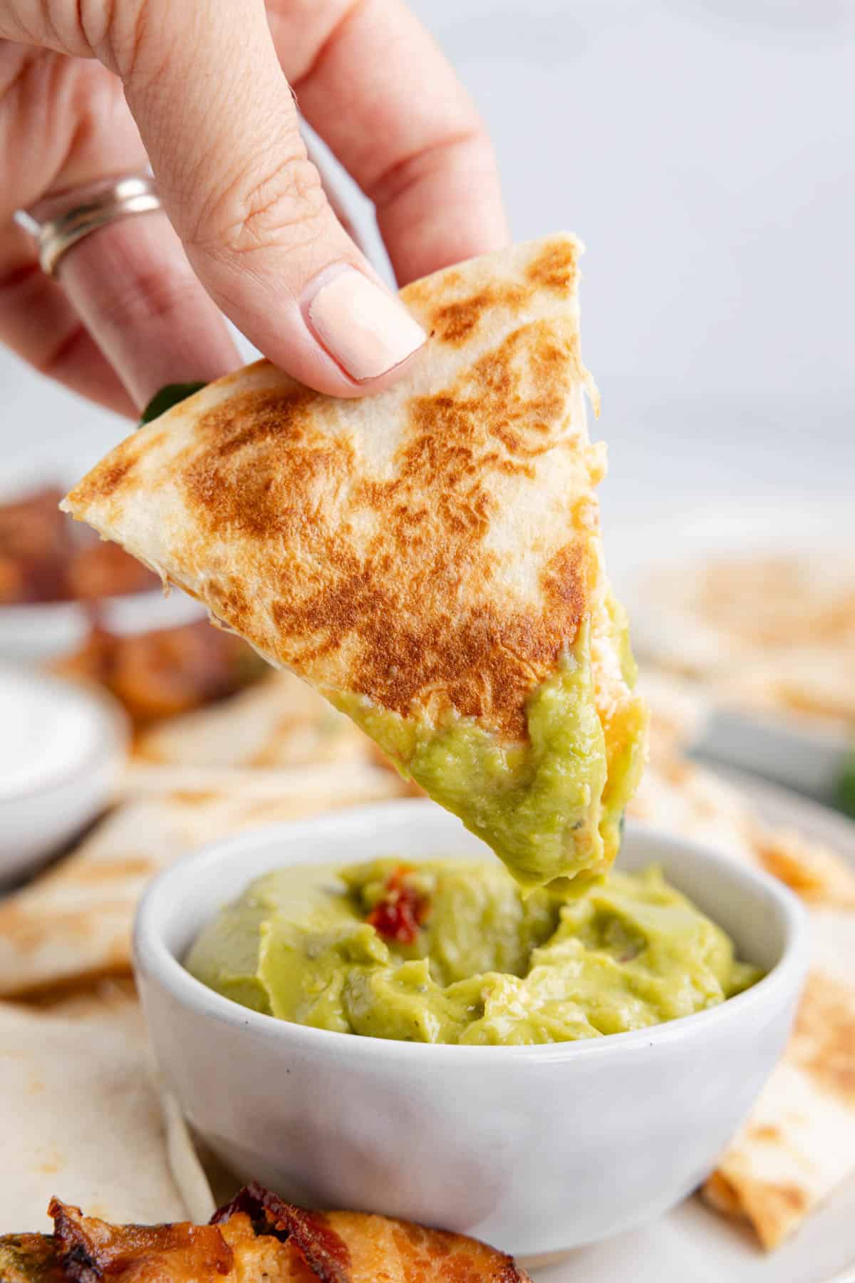 A hand holding a triangle of quesadilla to dip it into guacamole.