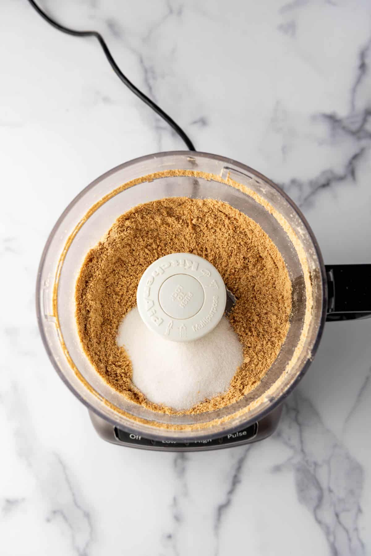 Combining crushed graham cracker crumbs and sugar in a food processor.