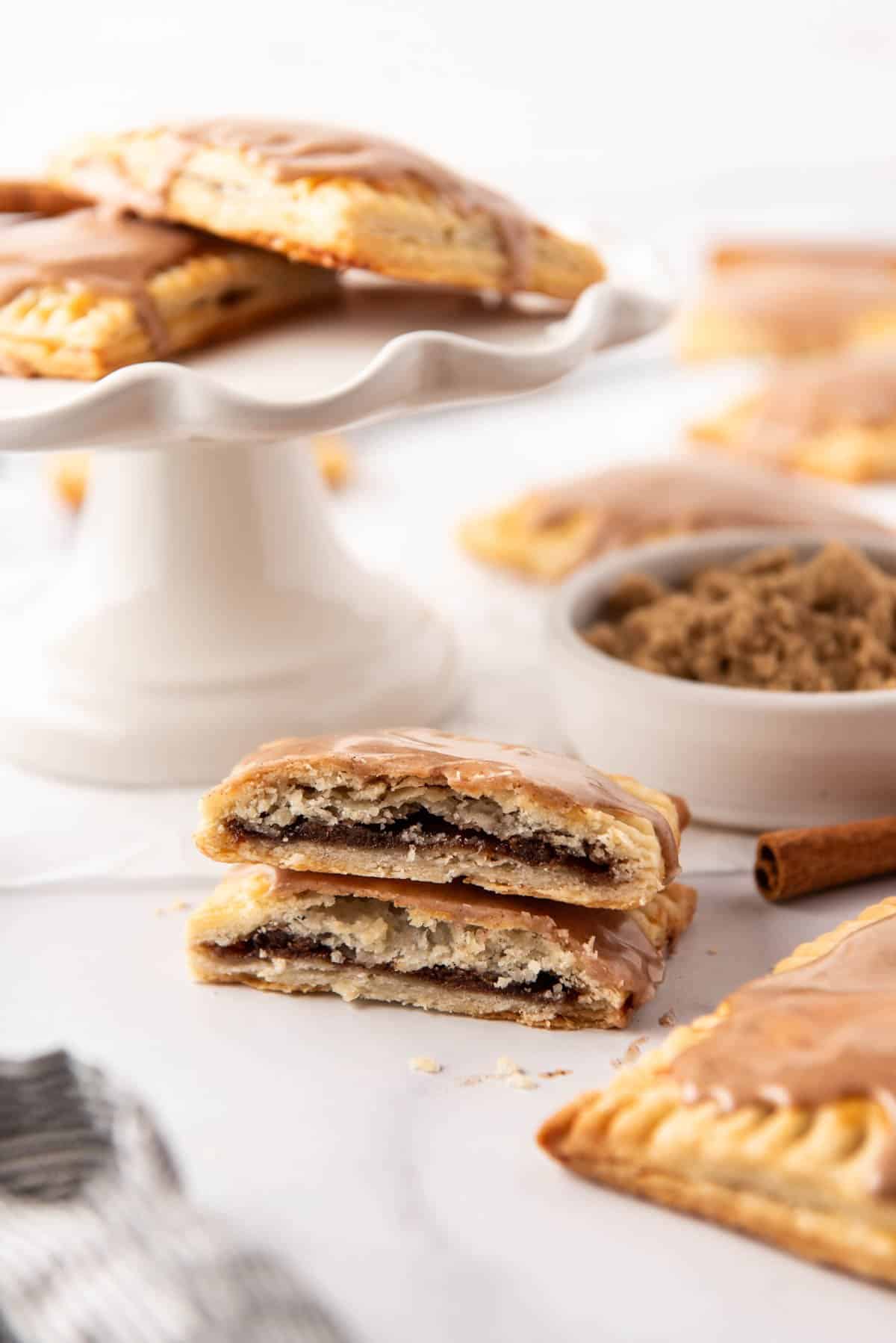 A brown sugar cinnamon pop tart that has been cut in half in front of a white cake stand with more pop tarts.
