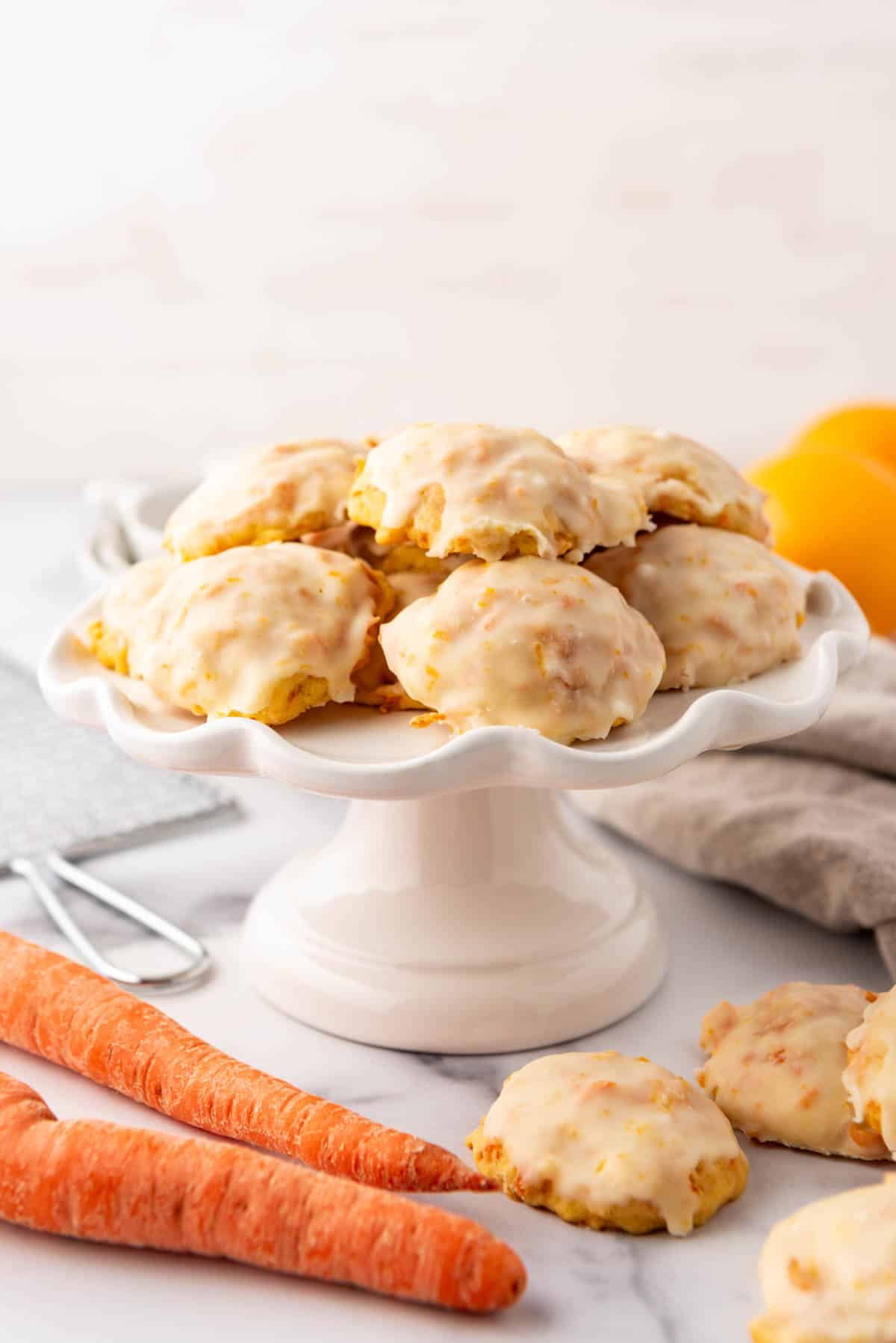 A white cake stand with orange glazed carrot cookies on top and carrots in front.