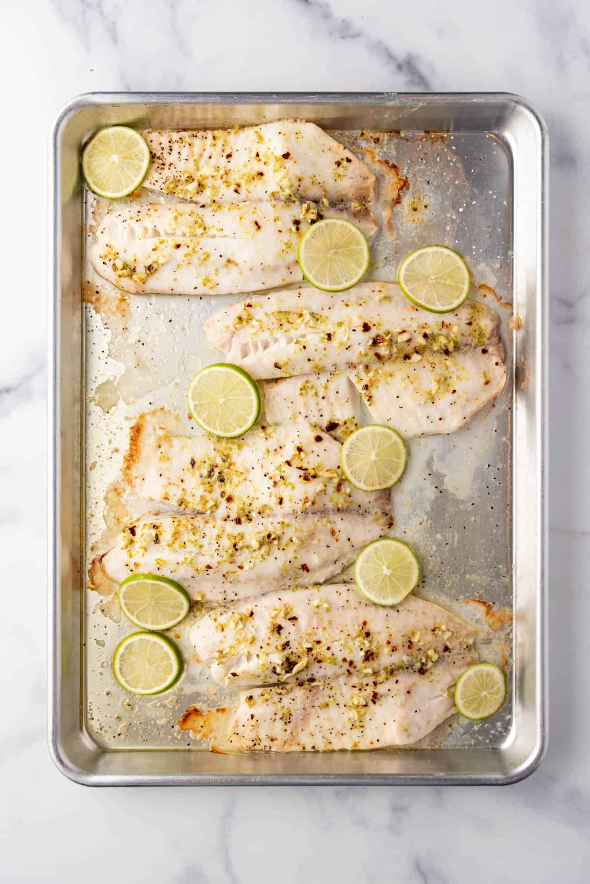 An overhead image of baked tilapia on a baking sheet with sliced limes on top.