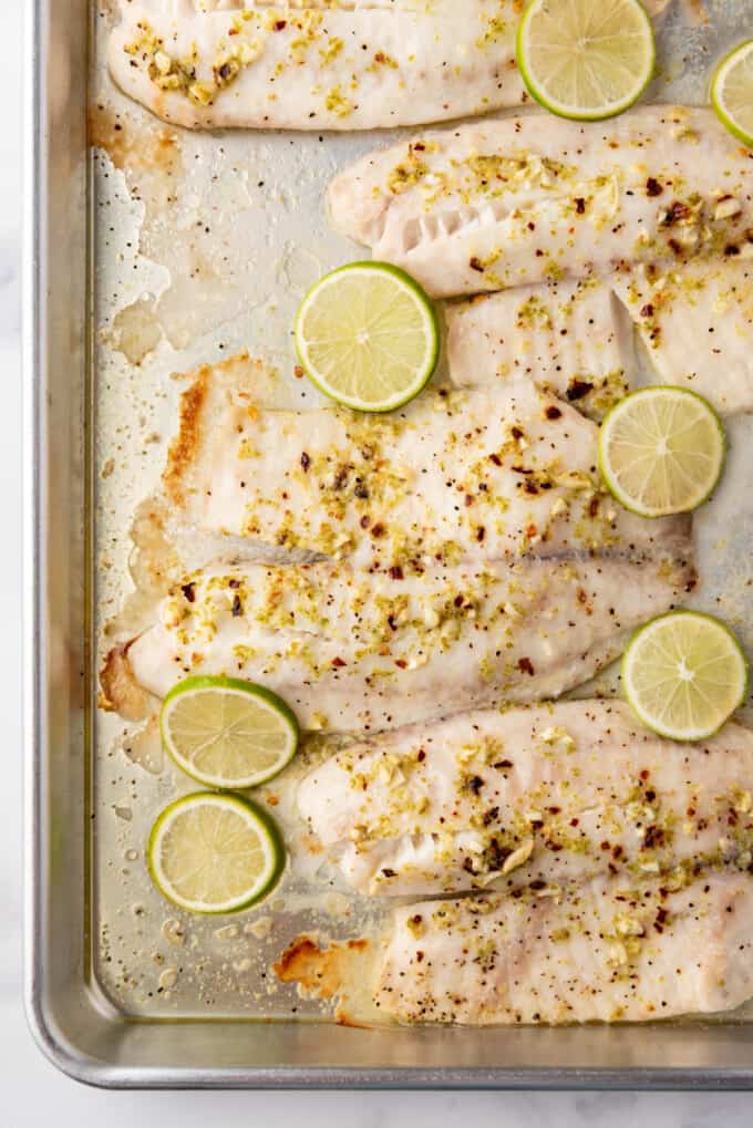 An overhead image of baked tilapia on a baking sheet with sliced limes on top.