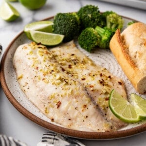 A piece of baked tilapia on a plate with lime wedges, steamed broccoli, and a piece of baguette.
