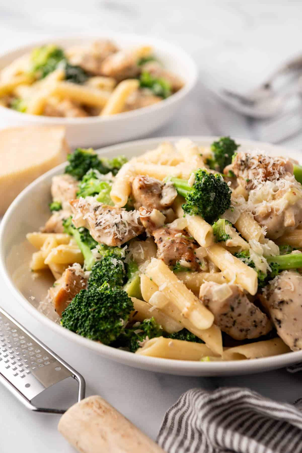 White pasta bowls filled with chicken broccoli pasta with creamy alfredo sauce next to a grater.