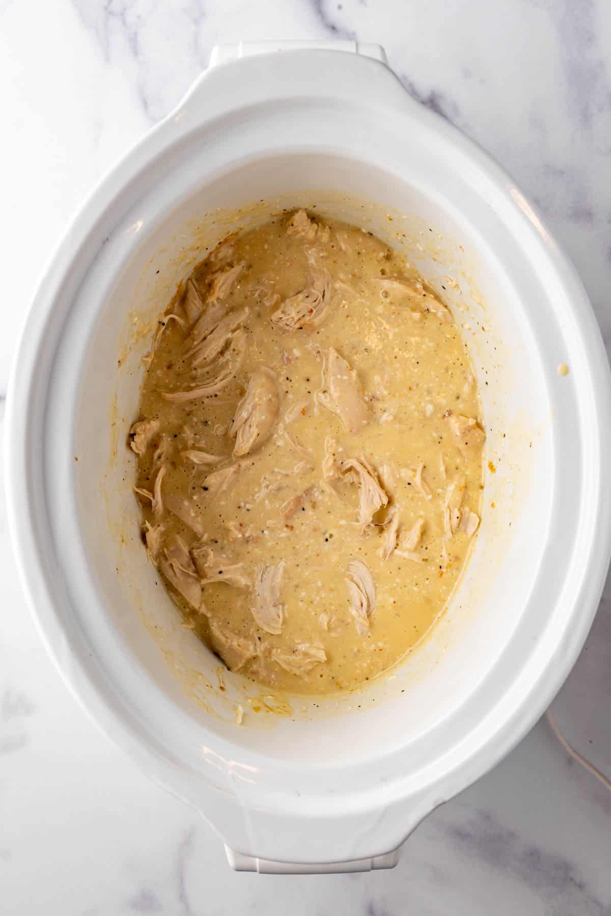 An overhead image of a white slow cooker with shredded chicken in a creamy sauce.