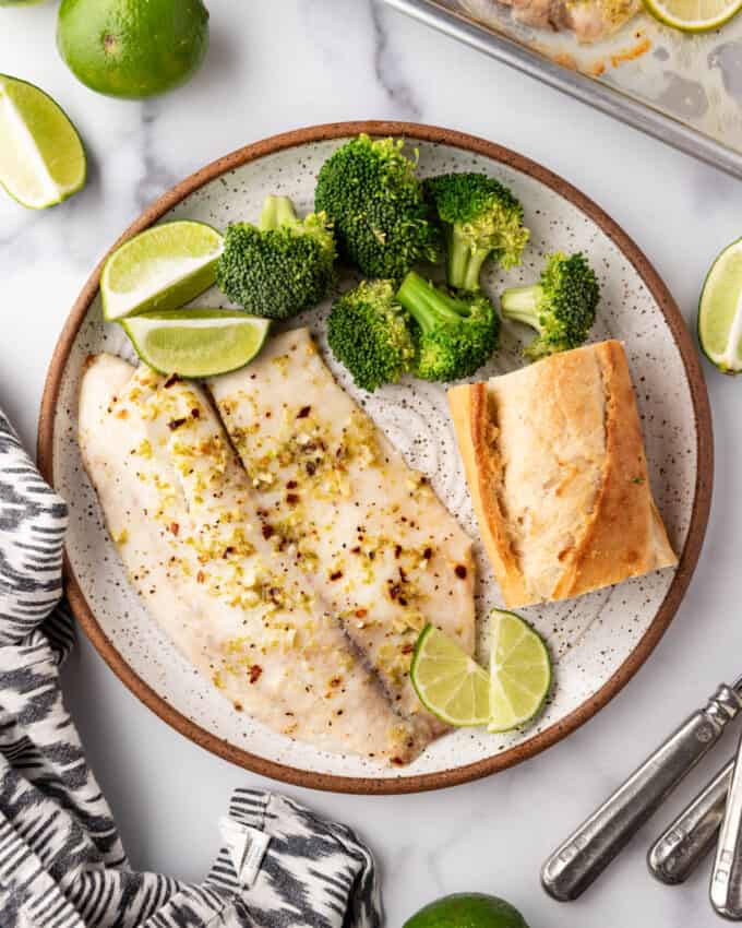 An overhead image of a plate of baked tilapia with steamed broccoli and a baguette.