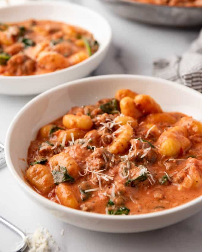 Pasta bowls of creamy sausage gnocchi in front of a pan of gnocchi in tomato sauce.