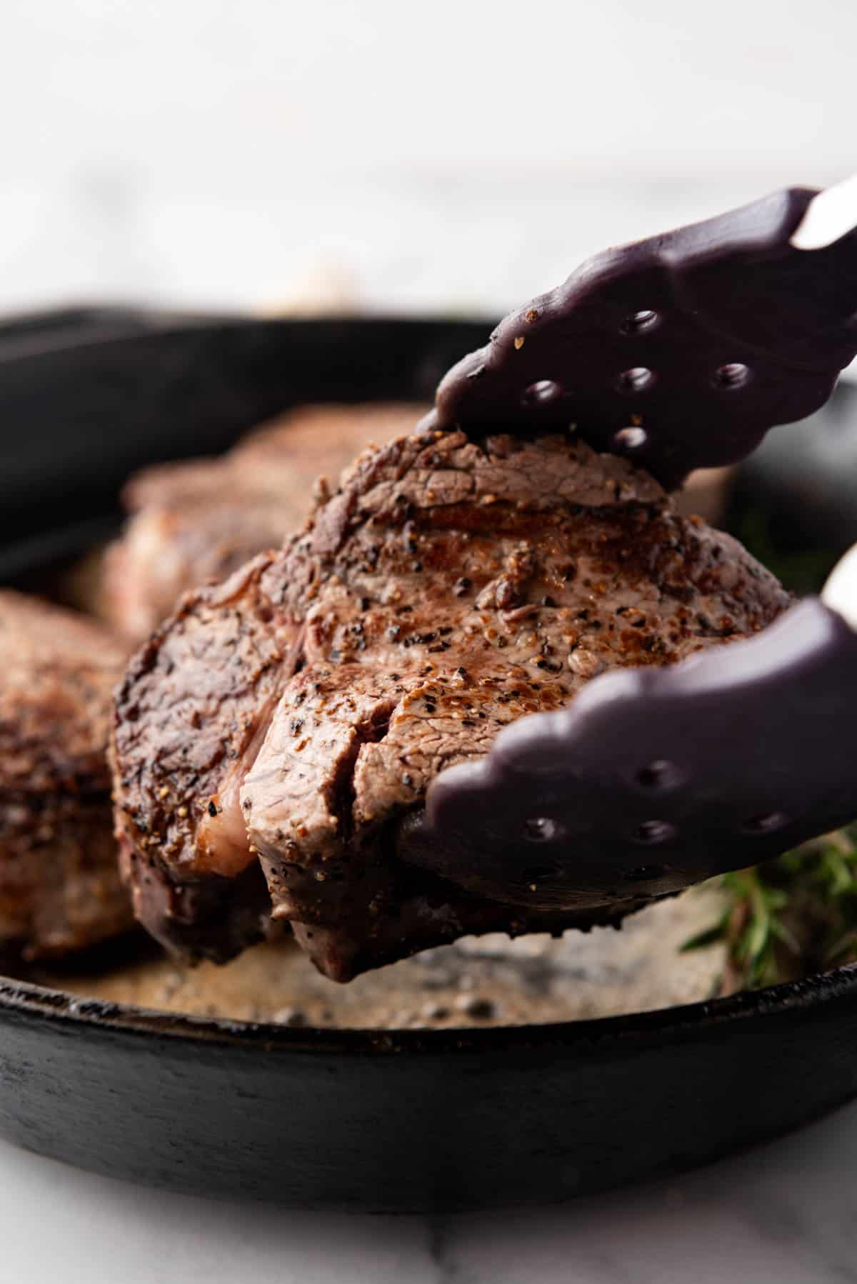 A filet mignon steak being lifted out of a cast iron skillet with tongs.