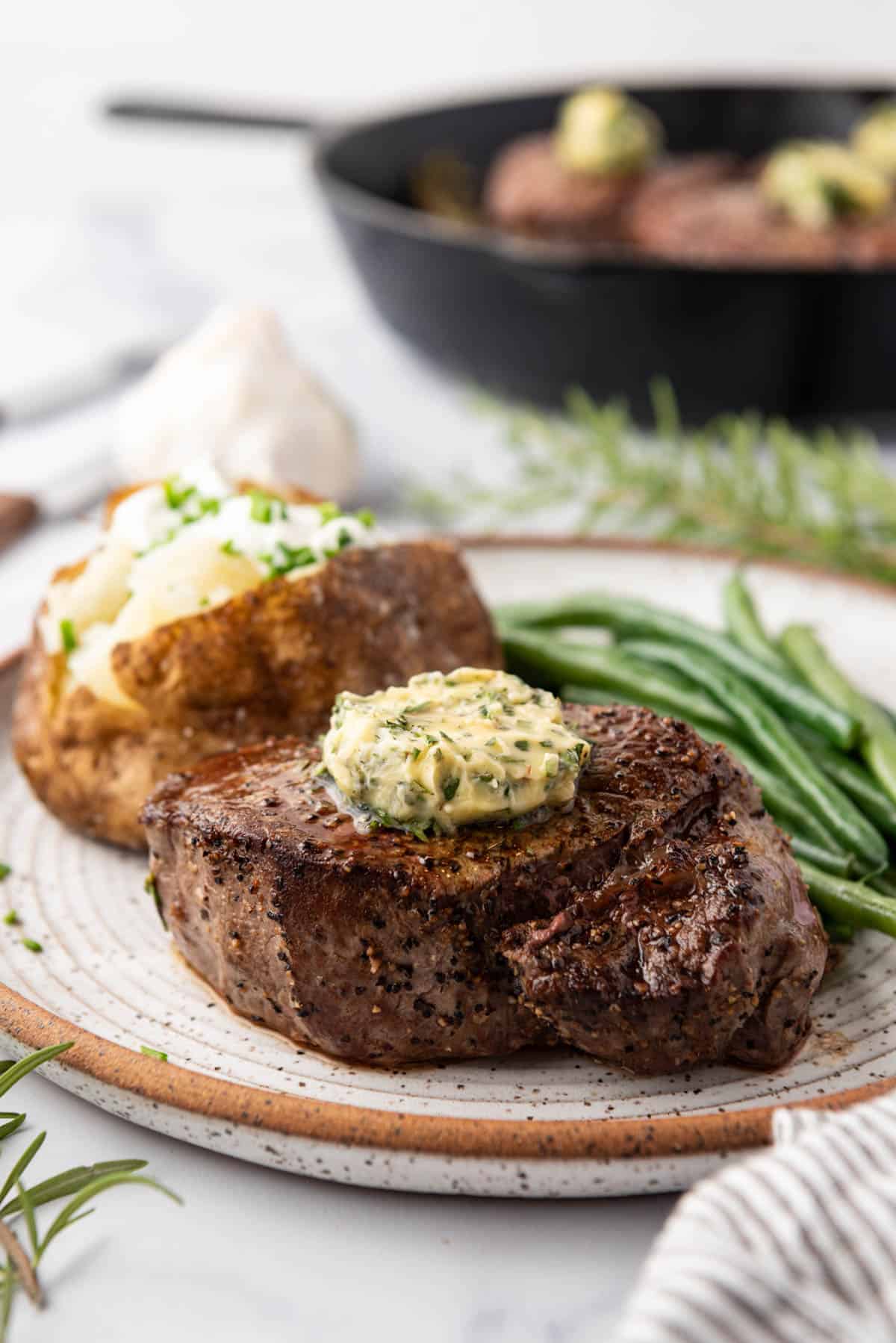 A cooked filet mignon on a plate with a baked potato, green beans, and garlic herb butter.
