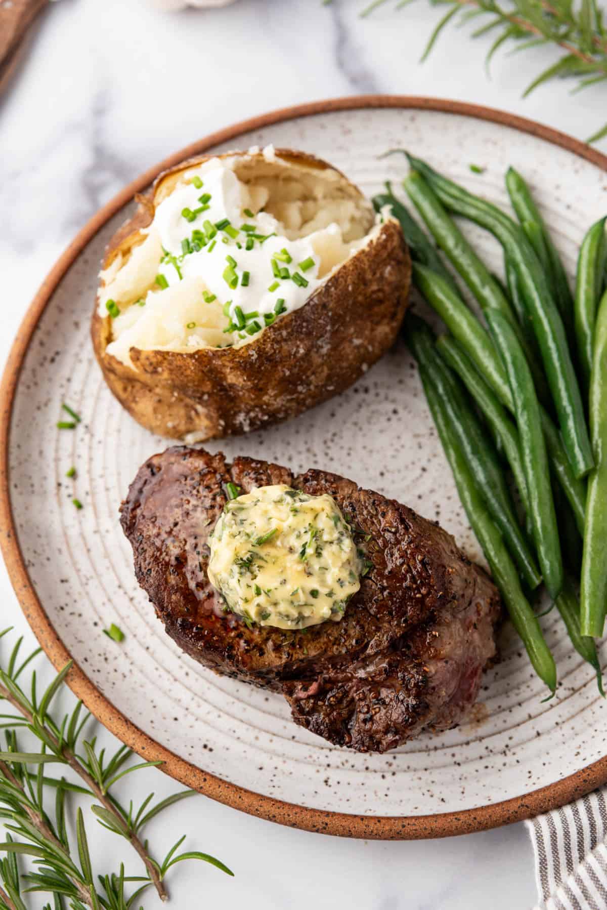 An overhead image of filet mignon on a plate with garlic herb butter, baked potato, and steamed green beans.