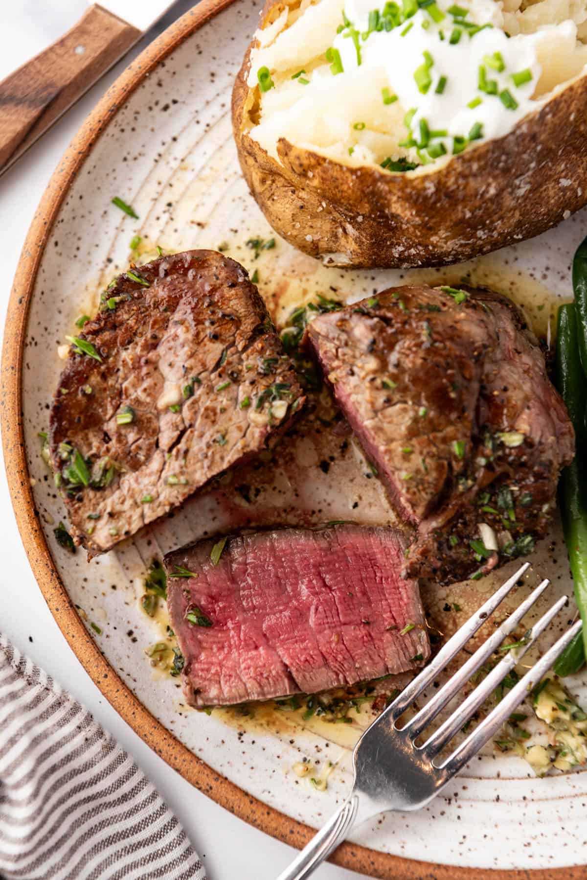 An overhead image of a filet mignon steak that has been cut in half with a slice of the medium-rare meat laying flat on the plate beside a baked potato and fork.