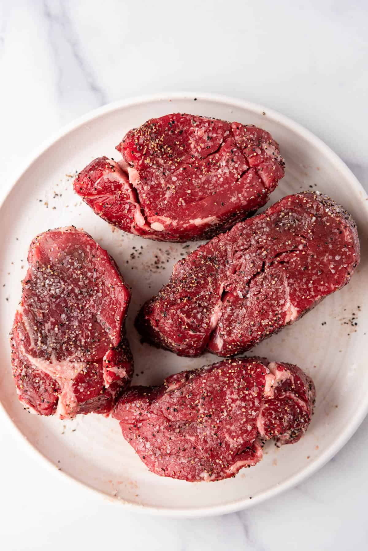Four filet mignon steaks seasoned with salt and pepper on a plate.