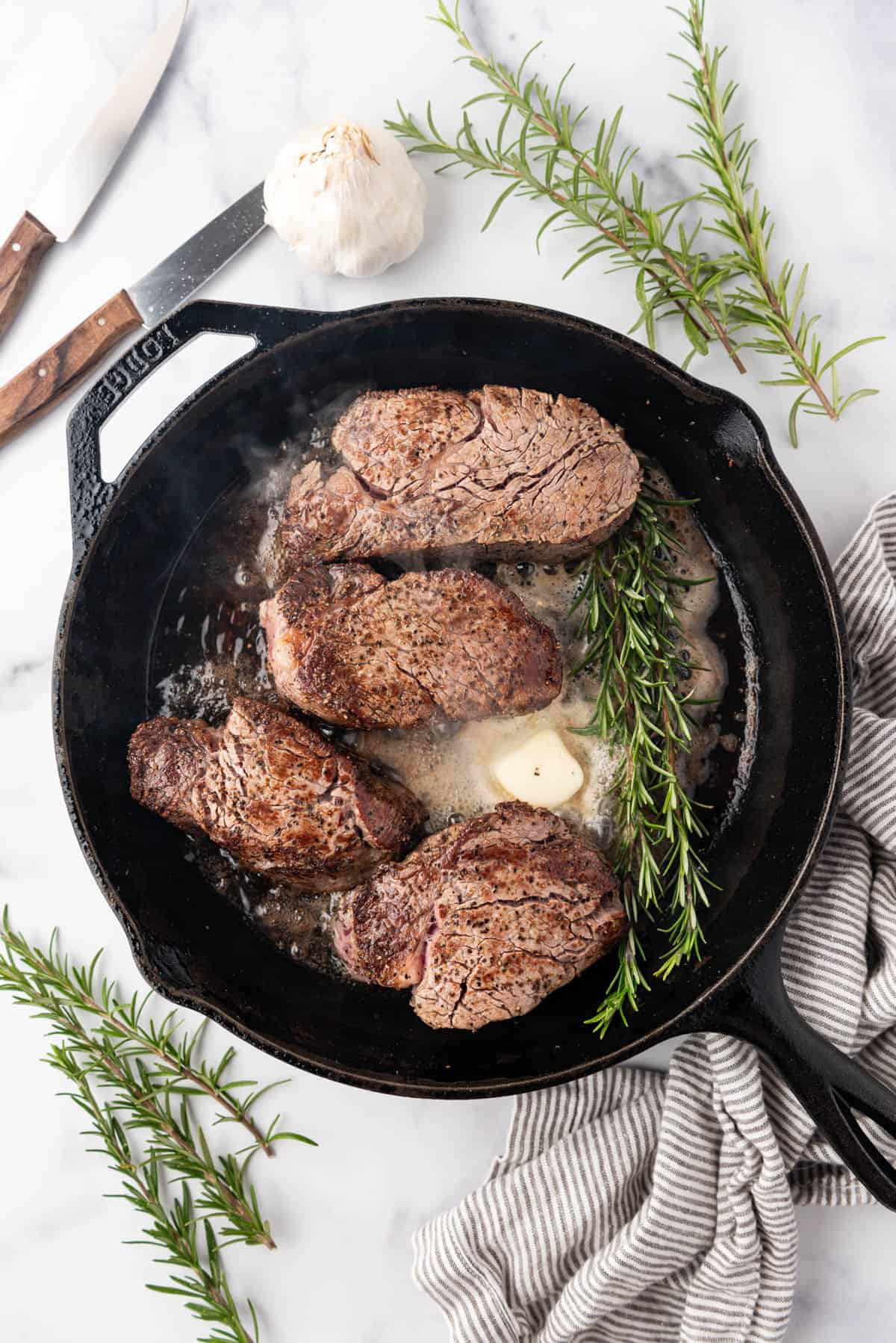 Adding butter and fresh rosemary to a cast iron skillet with filet mignon in it to finish the filet mignon.