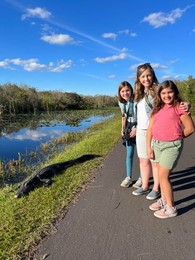 A mom and two daughters on a walking path next to an alligator in Everglades National Park at Shark Valley.
