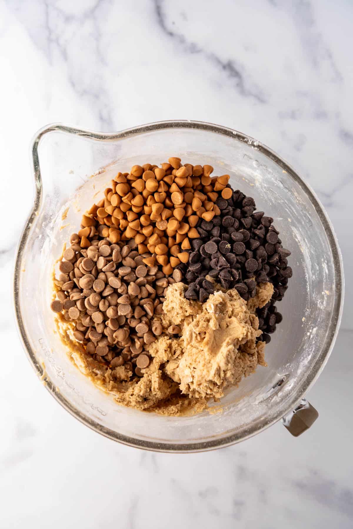 Adding chocolate chips, peanut butter chips, and butterscotch chips to peanut butter oatmeal cookie dough in a glass bowl.