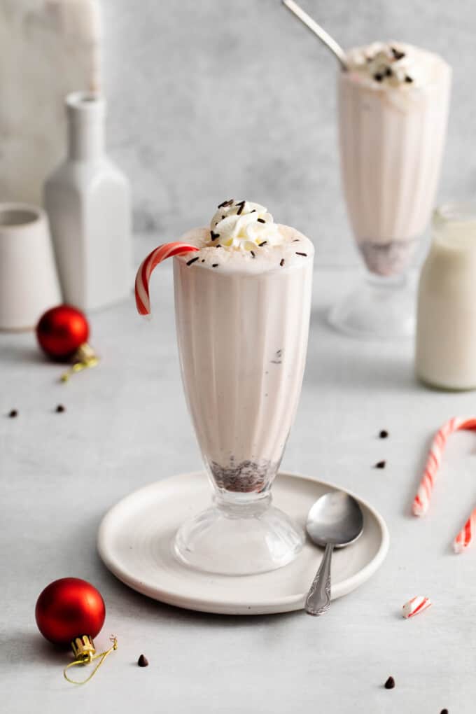 A homemade peppermint bark milkshake in a tall glass on a plate with scattered ornaments and candy canes nearby.