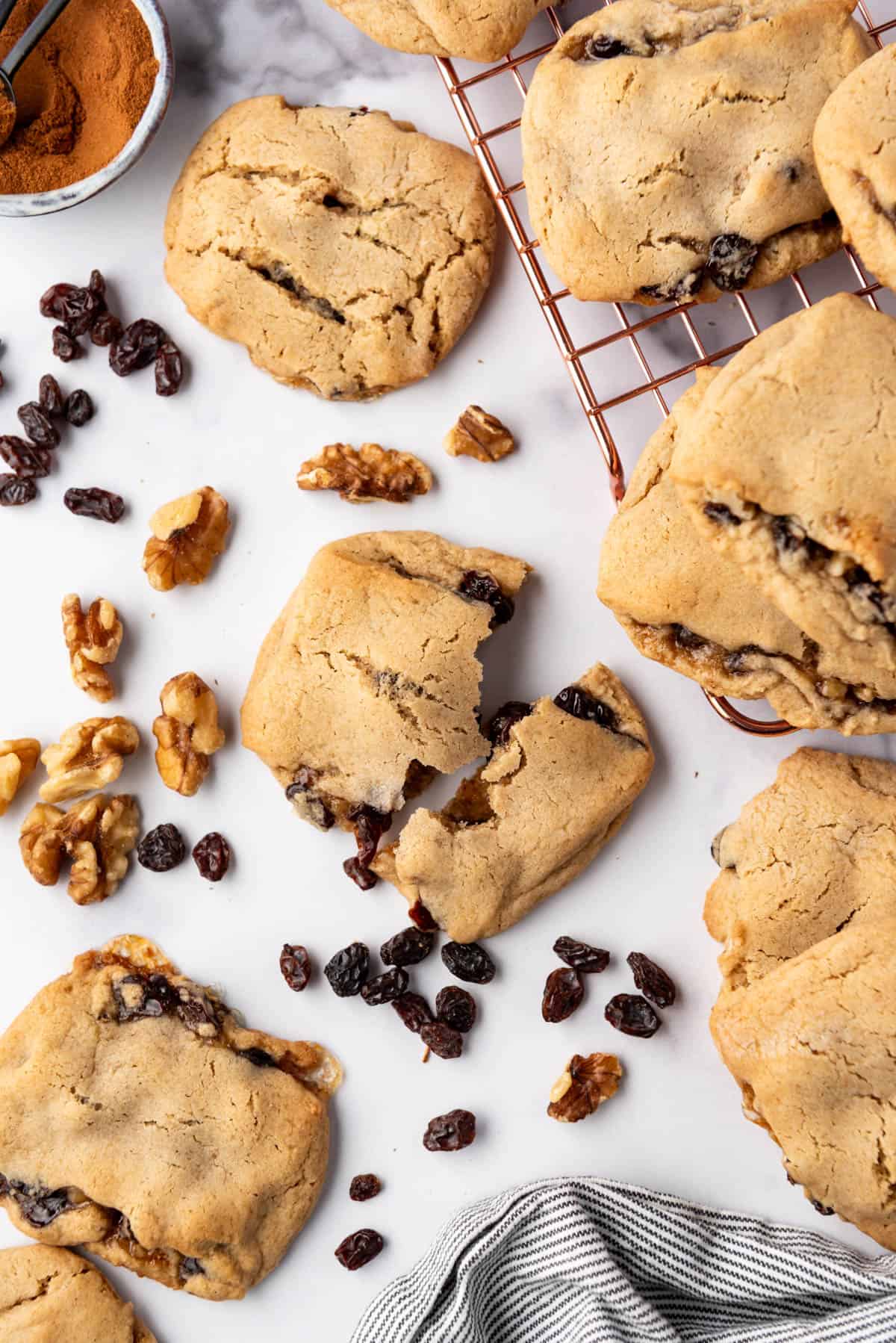 An overhead image of raisin-filled cookies with one of them broken in half with raisins and walnuts scattered around.
