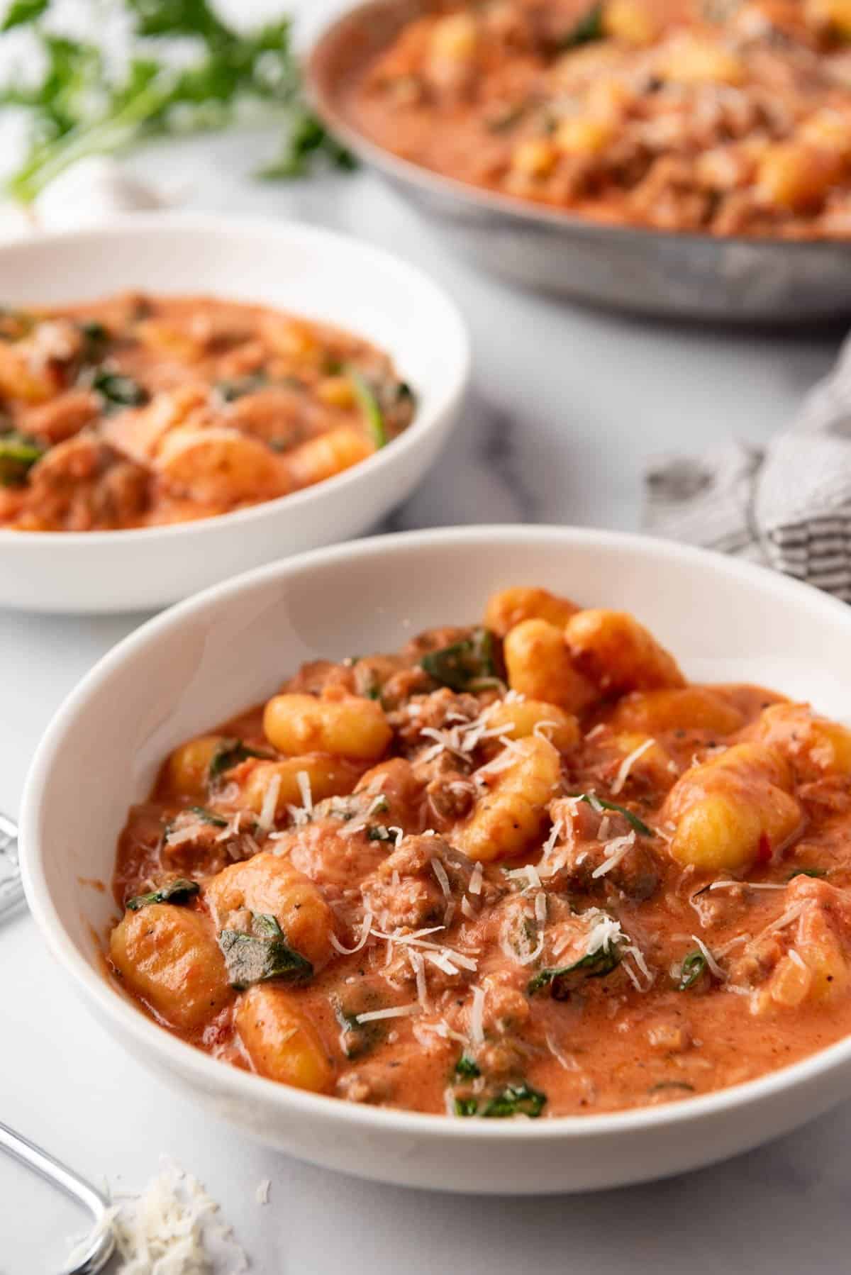 Pasta bowls of creamy sausage gnocchi in front of a pan of gnocchi in tomato sauce. 