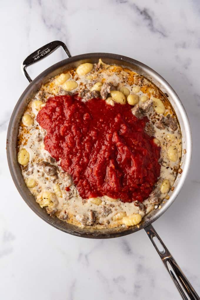 Adding crushed tomatoes to gnocchi and sausage in a large skillet.