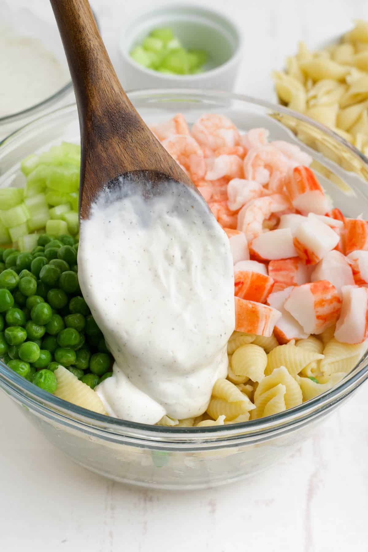Adding a creamy dressing to a mixing bowl filled with cooked shells pasta, crab, shrimp, and peas.