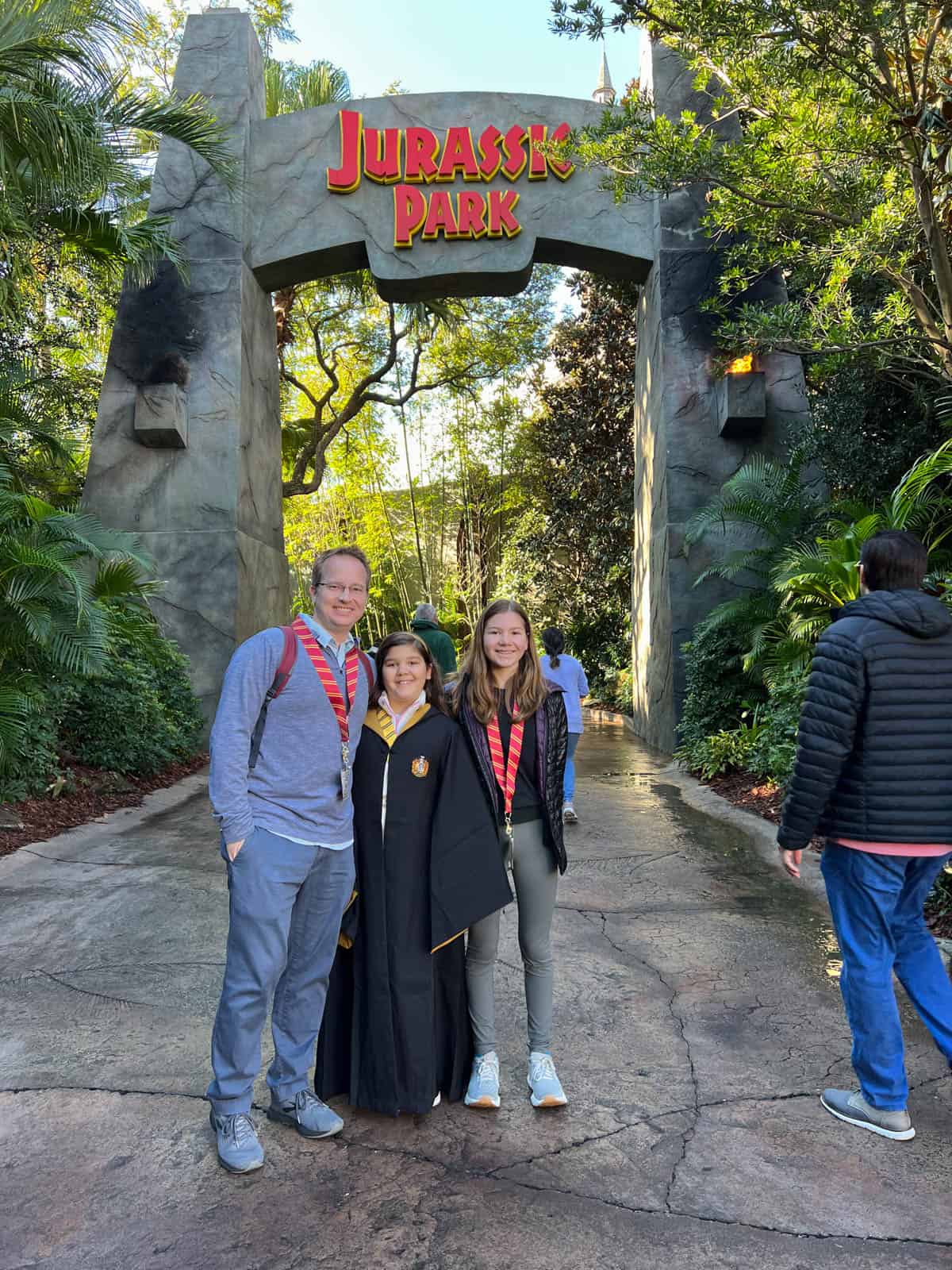 A dad and two daughters in front of the Jurassic Park sign at Universal Islands of Adventure theme park.