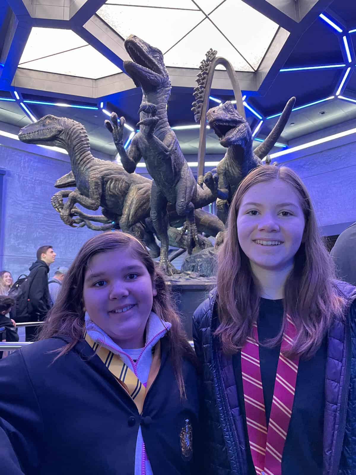 Two girls in front of a statue of raptors on the Velocicoaster ride.