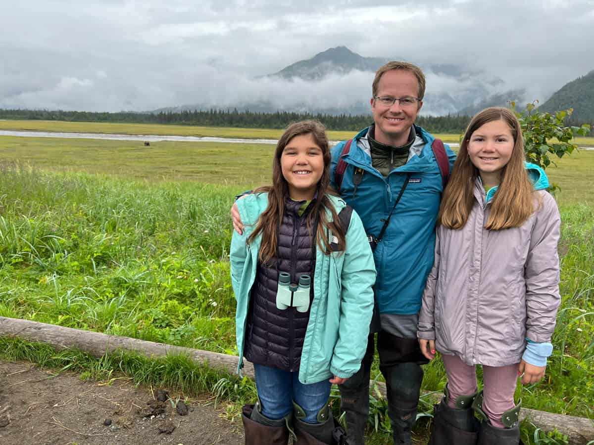 A dad and his two daughters in Alaska with bears in a meadow behind them.