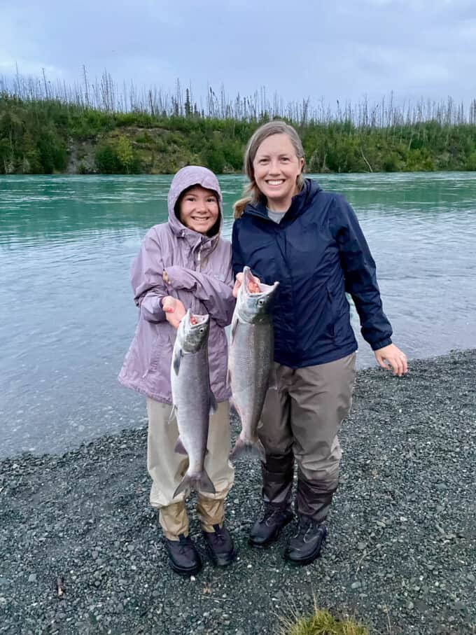A mom and her daughter each holding a sockeye salmon next to a river.
