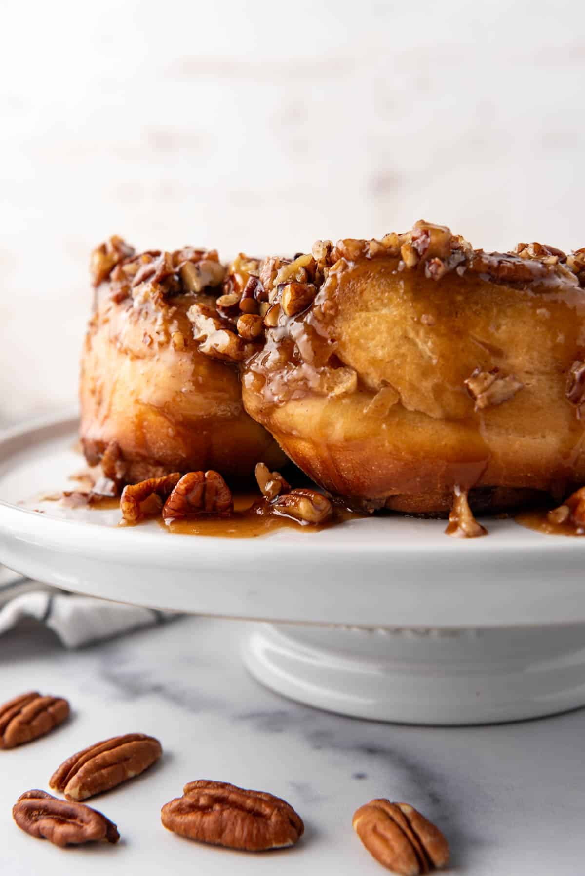 A close up image of a side view of caramel pecan sticky buns on a white cake stand.