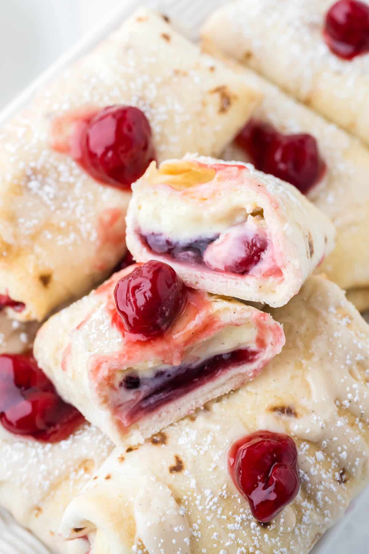A cheesecake chimichangas with cherry filling that has been sliced in half on top of more chimichangas.