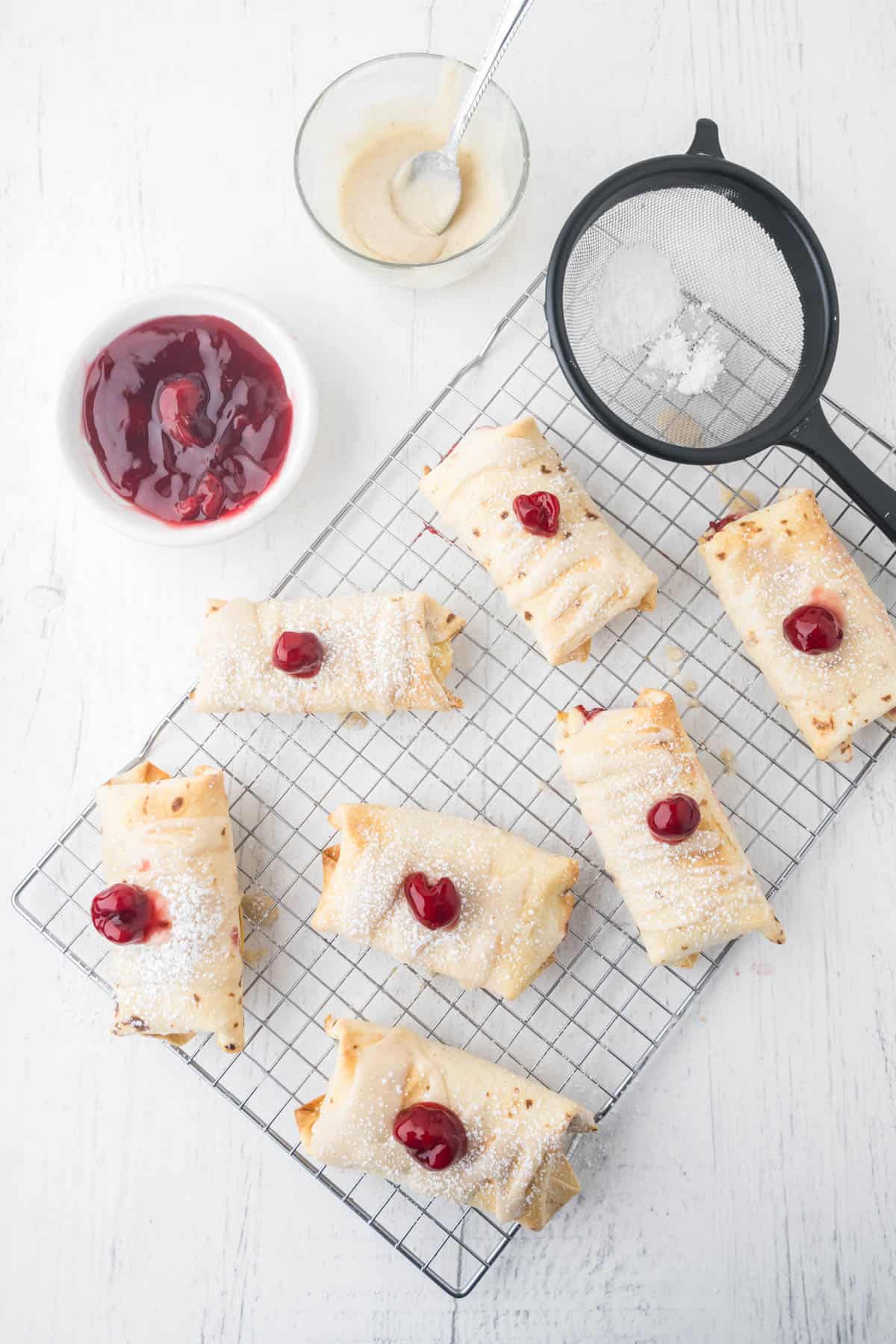 An overhead image of cheesecake chimichangas on a wire cooling rack decorated with cherry pie filling on top.