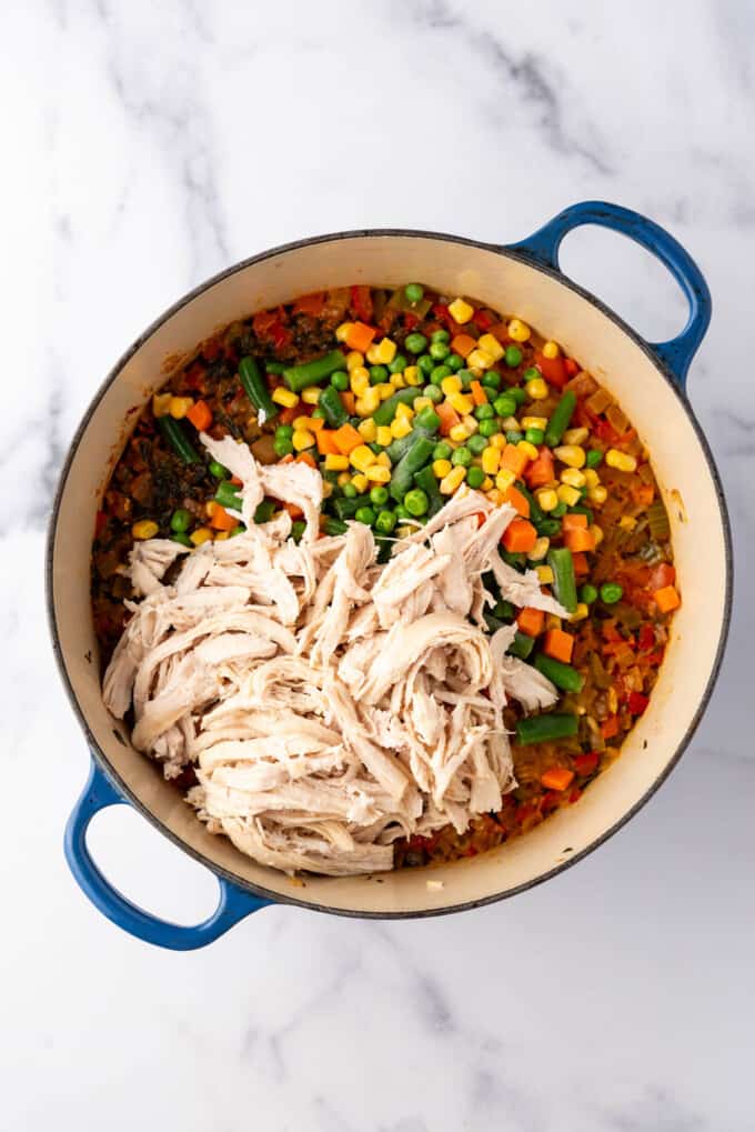 Adding shredded chicken and frozen mixed veggies to a large pot of arroz con pollo.