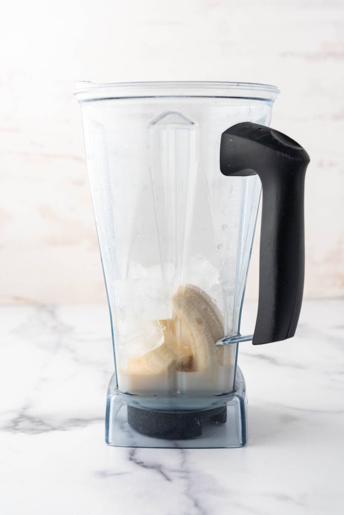 A large vitamix pitcher with milk, bananas, ice, sugar, and vanilla in it.