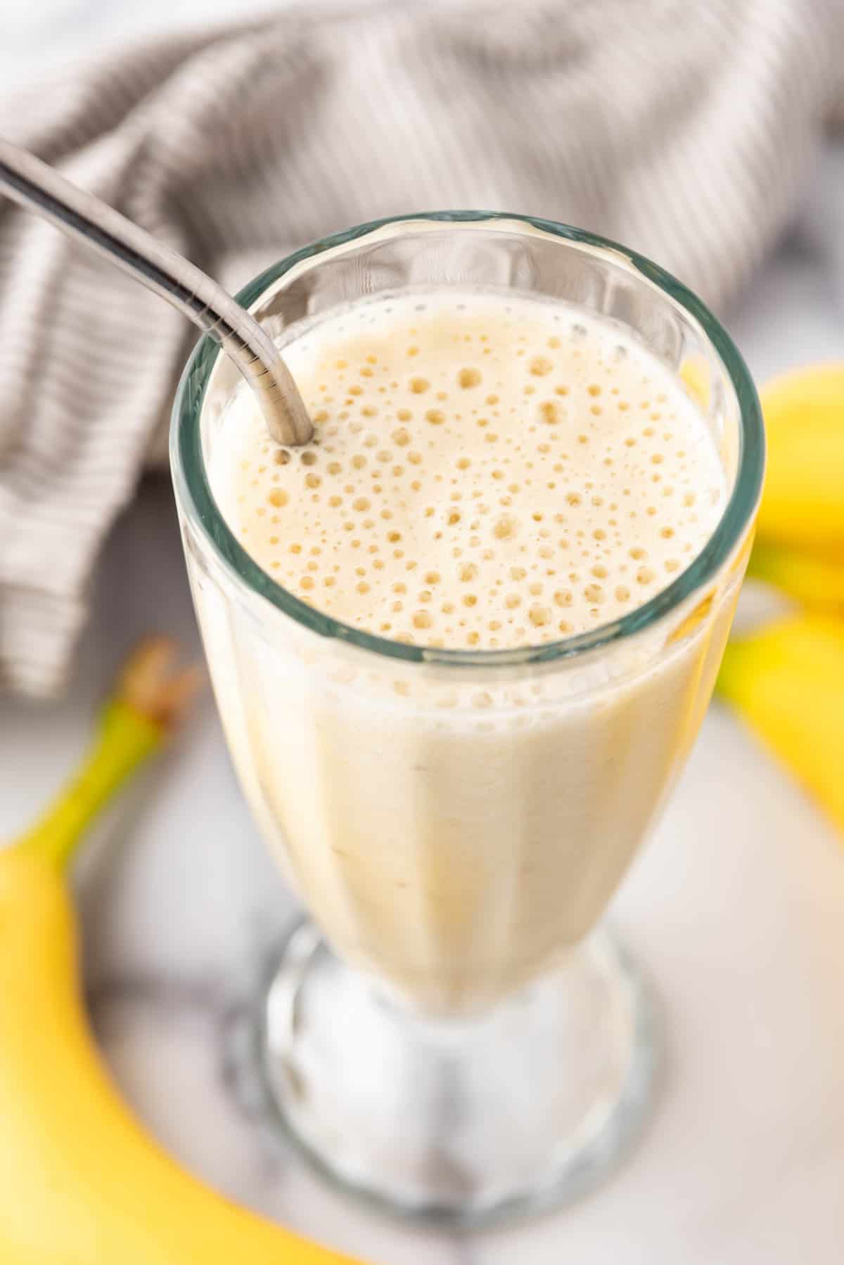 An overhead image of a blended banana smoothie with bubbles on top and a metal straw in it.