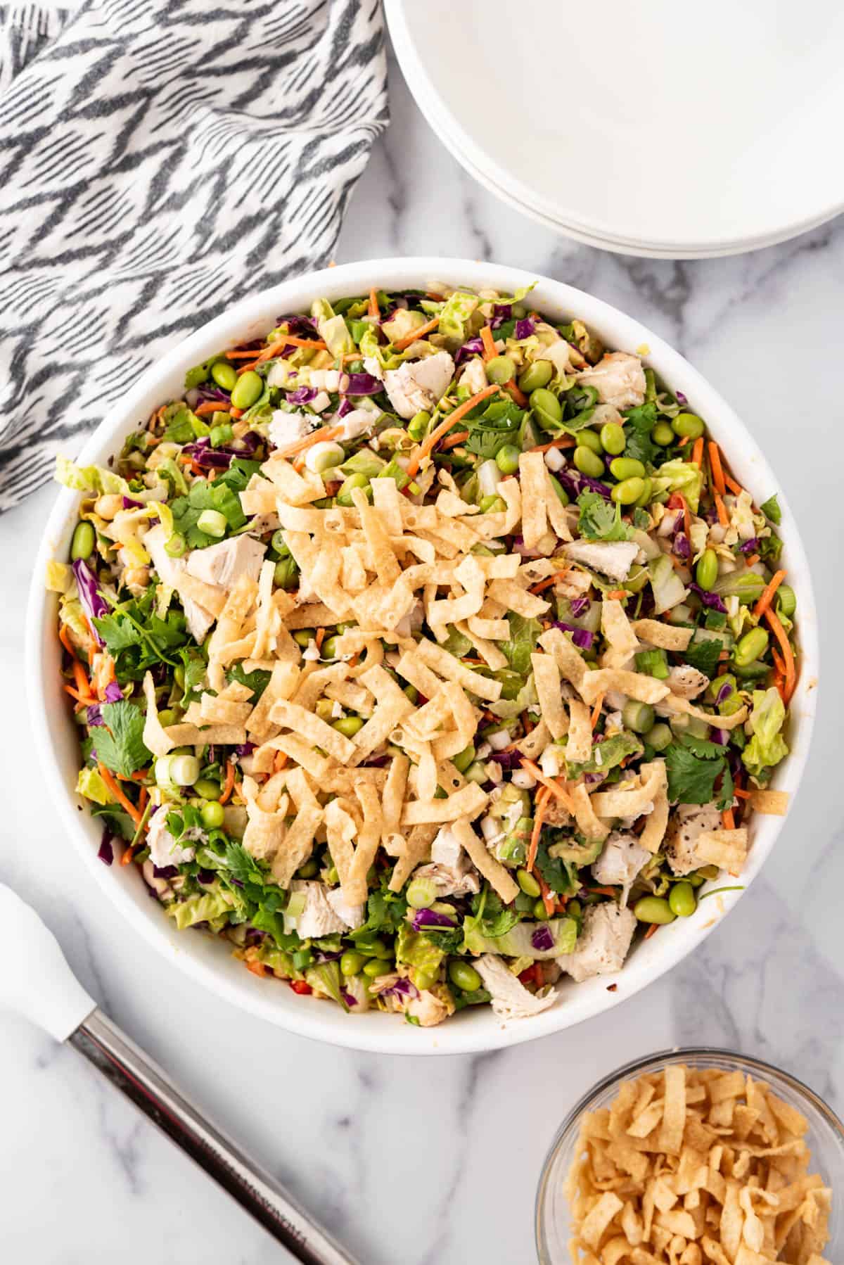 An overhead image of a big bowl of Thai-inspired chicken salad with a linen cloth and tongs next to it.