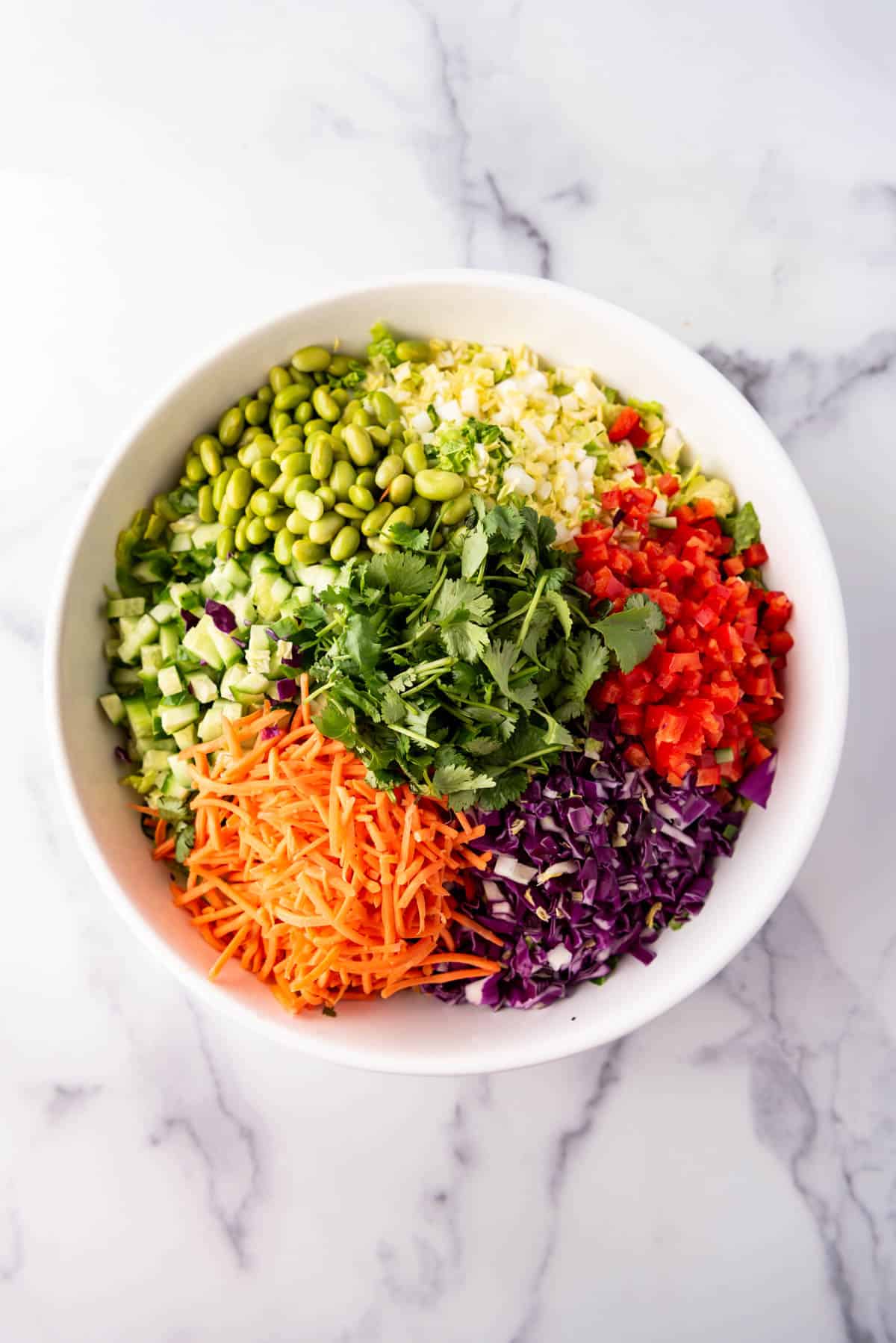 Combining colorful chopped vegetables like edamame, matchstick carrots, cilantro, red bell pepper, and cabbage in a large bowl.